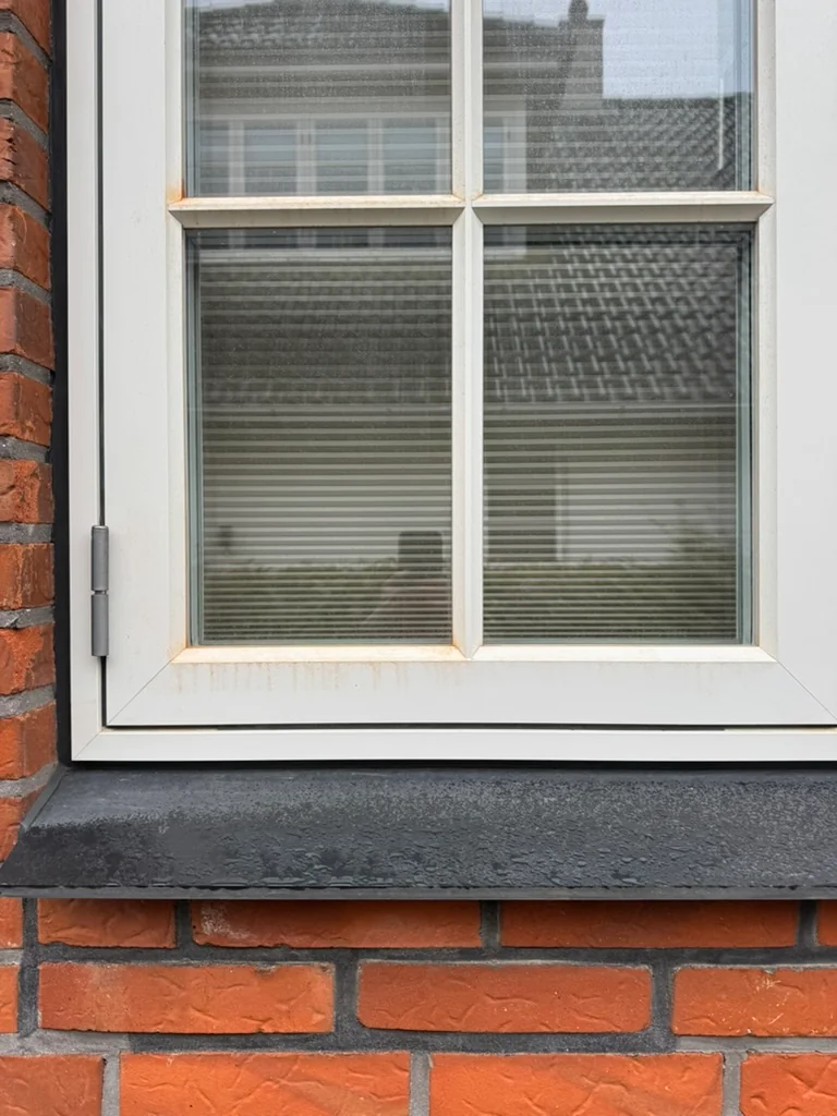 Rødt murstensstøv langs den hvide vinduesramme og sprosser før nedvaskning efter vinduesudskiftning.